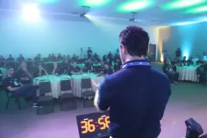 Palestrante conduz treinamento de gestão comercial para equipe em evento corporativo, com participantes atentos e cronômetro visível indicando dinâmica prática.