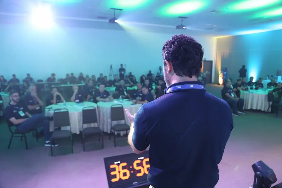 Palestrante conduz treinamento de gestão comercial para equipe em evento corporativo, com participantes atentos e cronômetro visível indicando dinâmica prática.